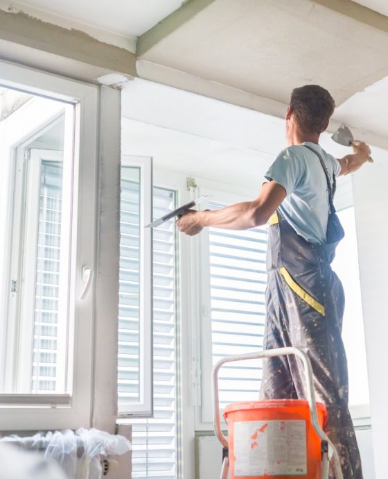 Thirty years old manual worker with wall plastering tools inside a house. Plasterer renovating indoor walls and ceilings with float and plaster.